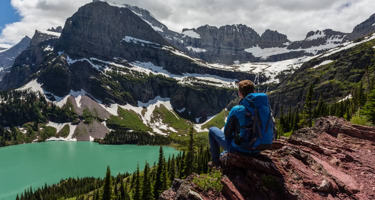 Backpacker with blue pack observes a turquoise alpine lake and jagged cliffs in Glacier National Park.