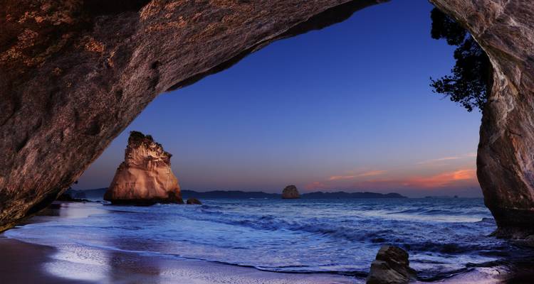 Dusk inside Cathedral Cove cave looking out to the sea and rock stacks bathed in deep blue hues.