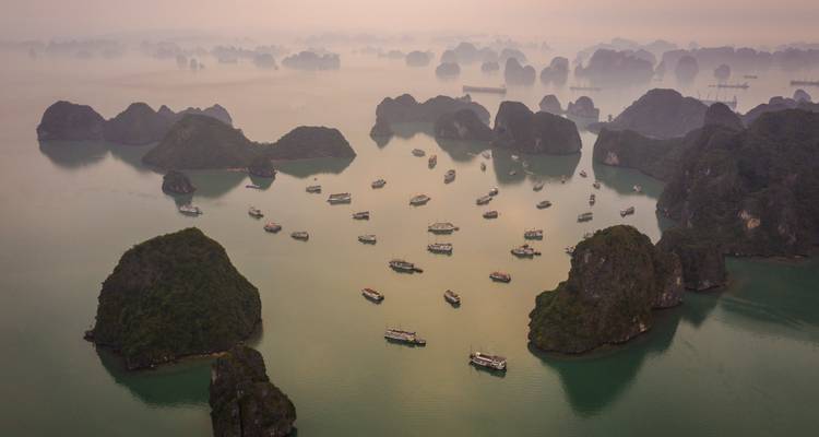 Luftaufnahme bei Sonnenaufgang mit Dutzenden von Kreuzfahrtschiffen, die zwischen Kalksteininseln in der nebligen Halong-Bucht verstreut sind.