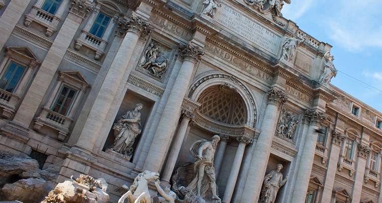 Primer plano arquitectónico en ángulo de la Fuente de Trevi de Roma con estatuas ornamentadas y columnas bajo un cielo azul.