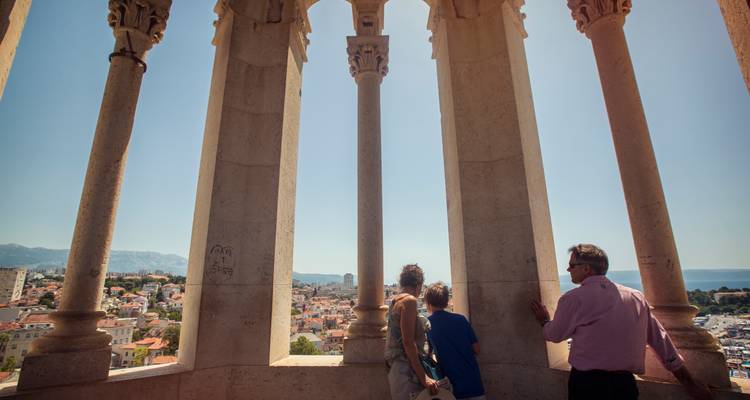 Una familia mira hacia afuera desde los arcos de un campanario de piedra sobre una ciudad costera croata y las montañas bajo una luz diurna brillante.