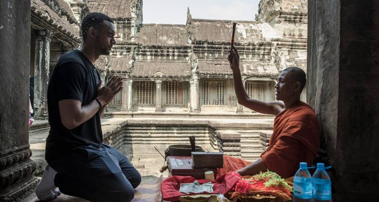 Viajero se arrodilla ante monje recibiendo ritual de bendición dentro de antiguo templo Angkor