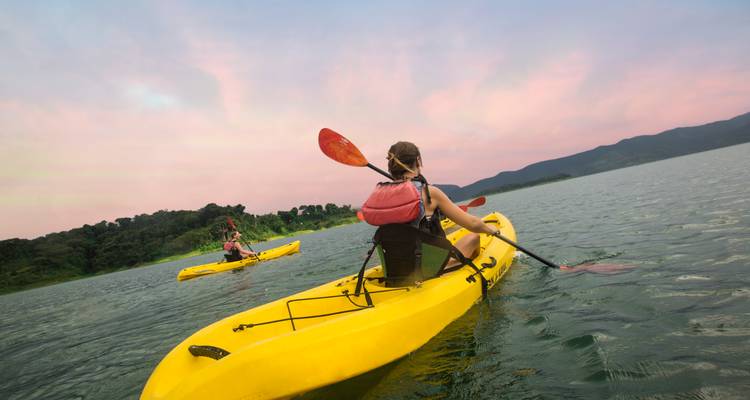 Los kayakistas reman botes amarillo brillante a través del tranquilo lago contra un cielo vespertino rosa pastel y colinas boscosas.