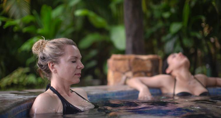 Dos mujeres se relajan tranquilamente en una piscina exterior cálida rodeada de exuberante vegetación tropical.