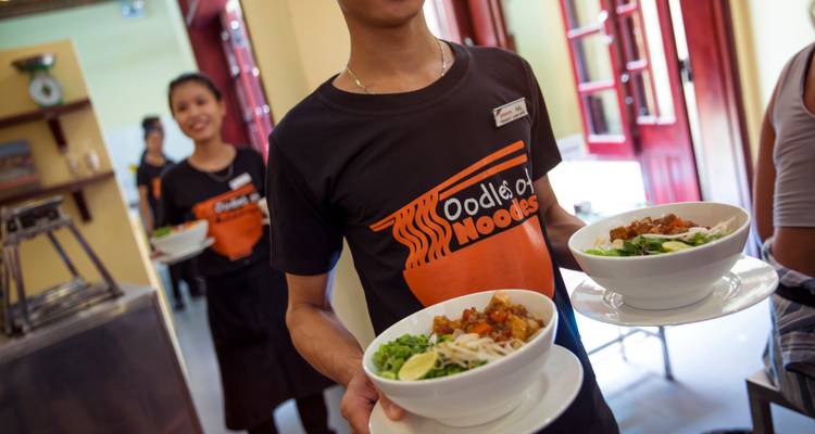 Mesero vistiendo una camiseta de 'Oodles of Noodles' carga tazones humeantes mientras otro miembro del personal sonríe detrás.