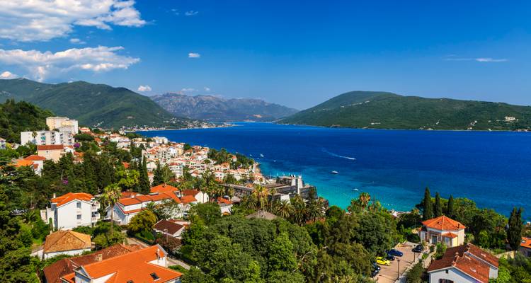 Vista panorámica de un pueblo costero con casas de techo rojo junto a una bahía azul profundo enmarcada por montañas verdes bajo un cielo despejado