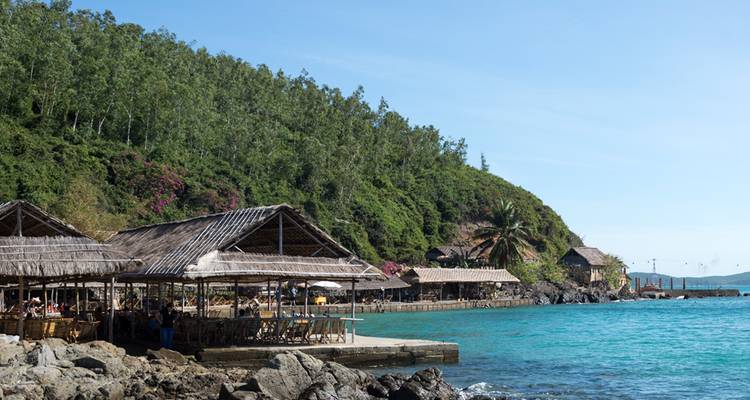 Cabanes en bois au bord de la mer au pied d'une colline boisée près d'une mer turquoise éclatante.