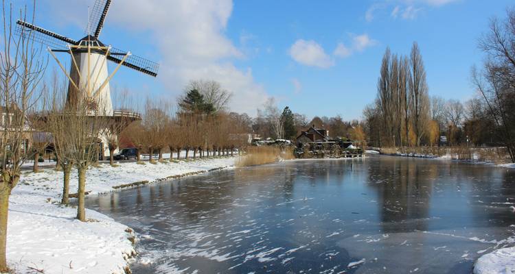 Traditionelle niederländische Windmühle neben teilweise gefrorenem Teich mit verschneiter Landschaft und blauem Himmel