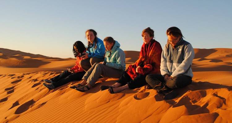 Fünf Menschen sitzen bei Sonnenaufgang auf einer Sanddüne.