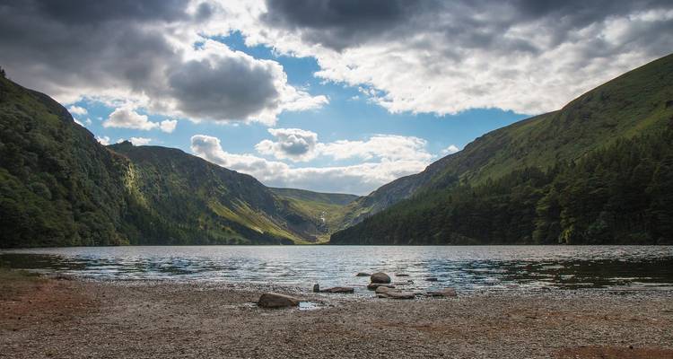 Lago de montaña enmarcado por pendientes boscosas empinadas bajo un cielo dramático con rayos de sol.
