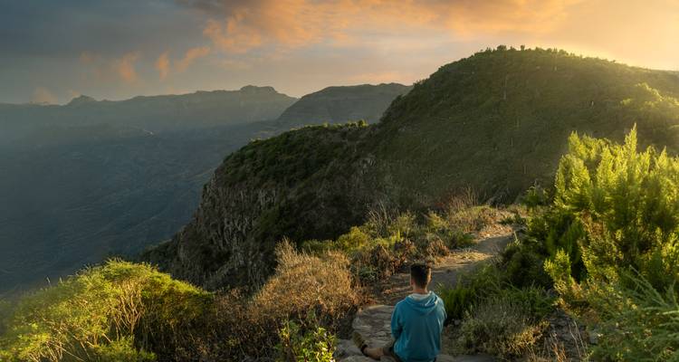Person sitting on a path at sunset overlooking hills and mountains.