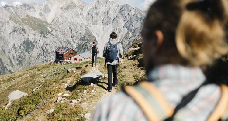 Wanderer, die auf eine Berghütte in alpiner Landschaft zugehen.