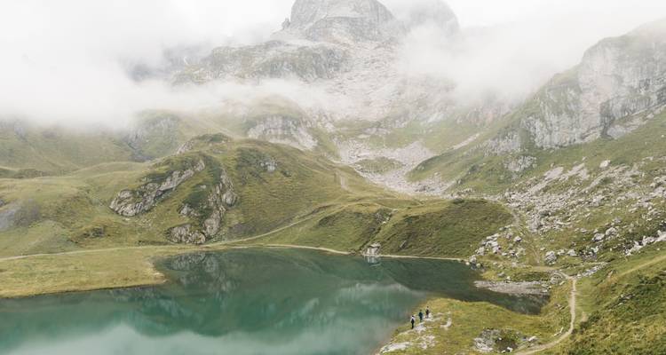 See mit Bergen im Nebel und Wanderern am Ufer.