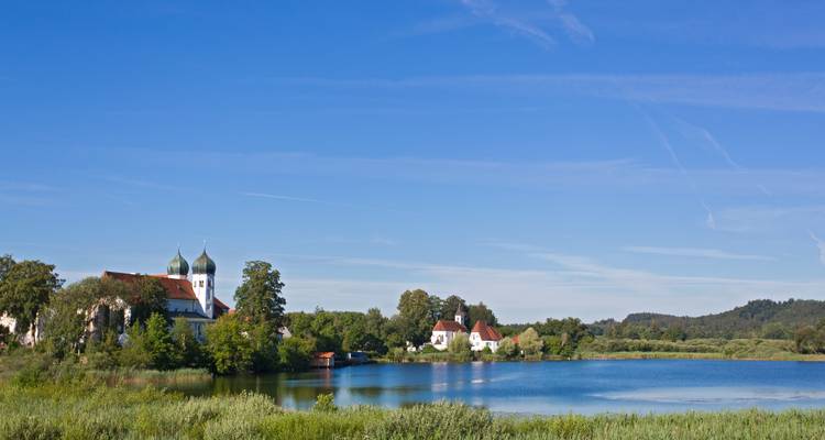 Petit village au bord du lac avec une église aux dômes en oignon se reflétant dans une eau calme sous un ciel bleu brillant.
