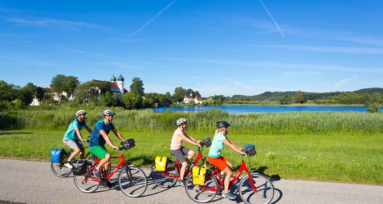 Quatre cyclistes chevauchent des vélos rouges le long d'un sentier pavé à côté d'un lac tranquille avec des bâtiments de village et des collines lointaines sous un ciel bleu lumineux.
