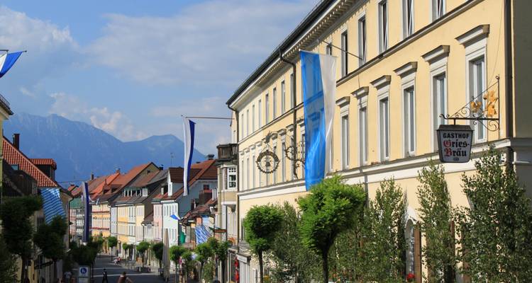 Charmante bayerische Straße gesäumt von pastellfarbenen Häusern und blauen Bannern, die zu den fernen Alpen führt.