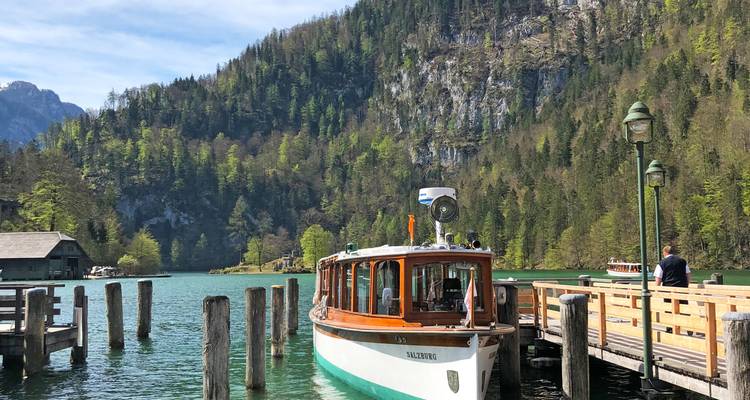 Passagierboot an einem Dock mit grünen Hügeln und Wald im Hintergrund.