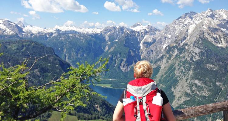 Ein Rucksacktourist mit einem roten Rucksack blickt über einen tiefen Alpensee, umgeben von schneebedeckten österreichischen Gipfeln.
