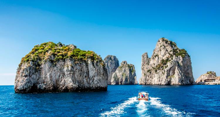 La lancha rápida deja un rastro blanco mientras se acerca a los imponentes Faraglioni de Capri bajo cielos azul brillante.