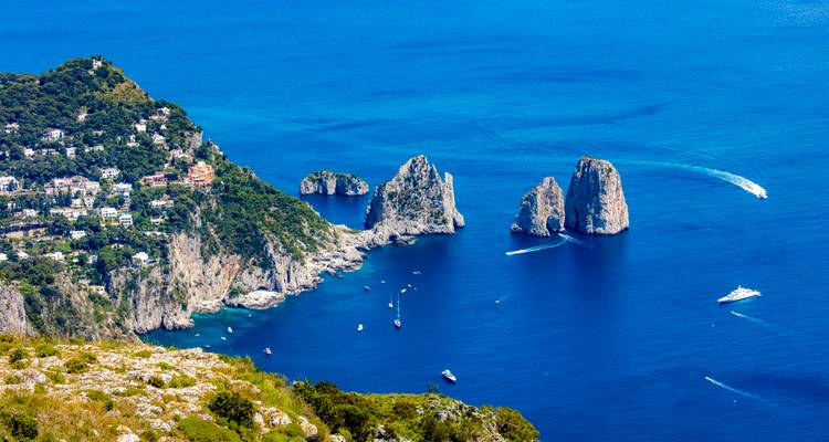 Vista elevada hacia las rocas Faraglioni en Capri con barcos trazando sendas blancas en las aguas azul profundo.