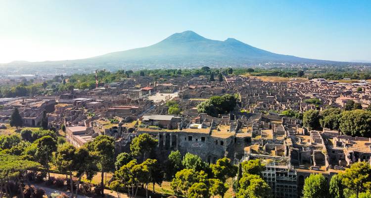 Vista aérea impresionante de las ruinas expansivas de Pompeya con el Monte Vesubio asomándose al fondo bajo un cielo despejado.