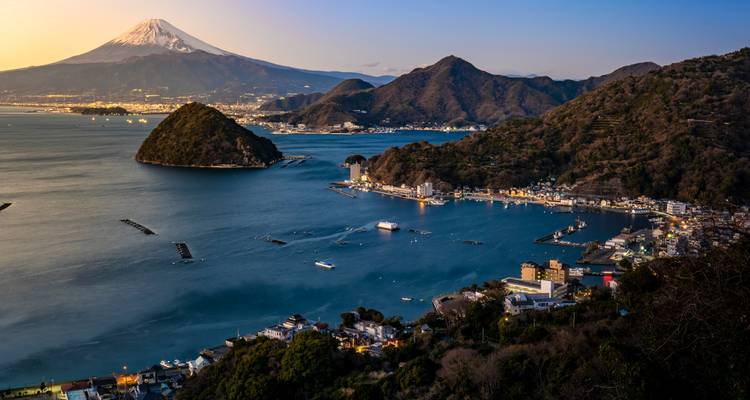 Dämmerungsblick auf den Berg Fuji, der sich über eine Küstenstadt und eine ruhige Bucht mit Fischernetzen erhebt