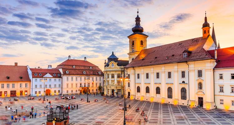 Der historische Hauptplatz von Sibiu, getaucht in warmes Sonnenuntergangslicht, mit farbenfrohen Barockgebäuden und vielen Menschen, die über den gemusterten Platz spazieren.