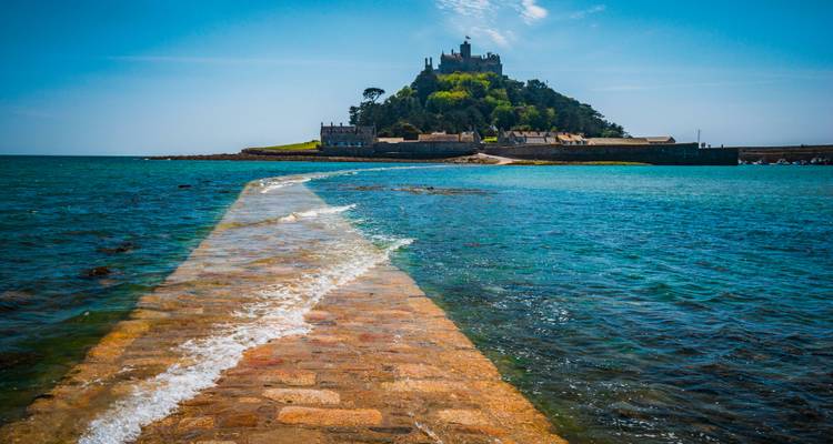 A stone causeway leads across sparkling blue water to Saint Michael's Mount crowned by a castle on a sunny day.