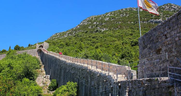 Long defensive stone wall snakes up a green hillside towards an arched gate beneath a cloudless sky
