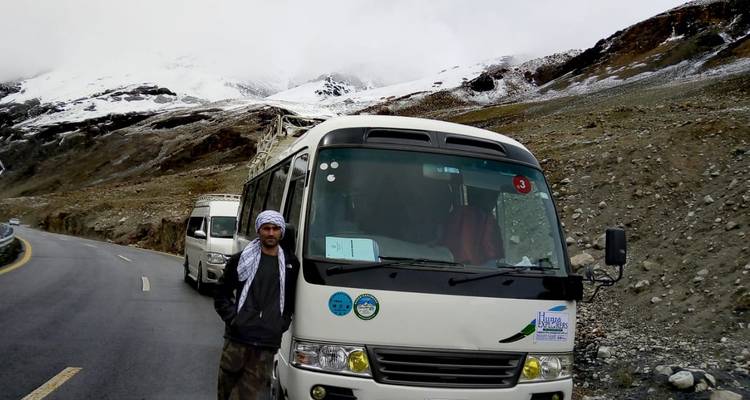 Ein Mann, der neben einem Touristenbus auf einer Straße steht, mit schneebedeckten Bergen im Hintergrund.