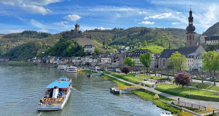 Flussuferblick auf Cochem mit Burg, Kirchturm und Ausflugsboot unter blauem Himmel.