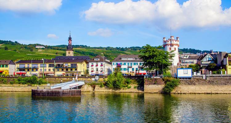 Small Rhine Valley town with church tower and riverside promenade under scattered clouds.