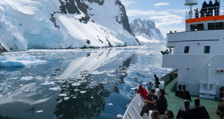Barco de crucero navegando por un canal angosto en la Antártida rodeado de montañas cubiertas de nieve y hielo flotante, con pasajeros observando desde la cubierta