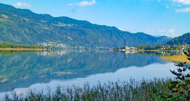 Der spiegelglatte See reflektiert die umliegenden grünen Hügel und eine kleine Seestadt unter einem blauen Sommerhimmel.