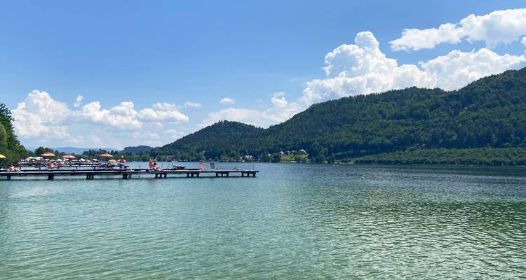 Ein Freizeitsee mit hölzernen Stegen und Schwimmern vor bewaldeten Hügeln unter aufgetürmten Wolken.