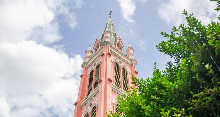 Roze kerktoren met een kruis, omlijst door weelderig groen gebladerte.