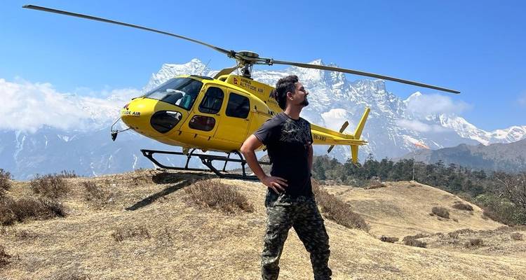 Man posing in front of a yellow helicopter on a mountain landscape.