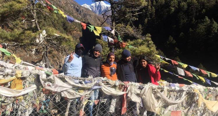 Group posing on a bridge with prayer flags, mountains in the background.