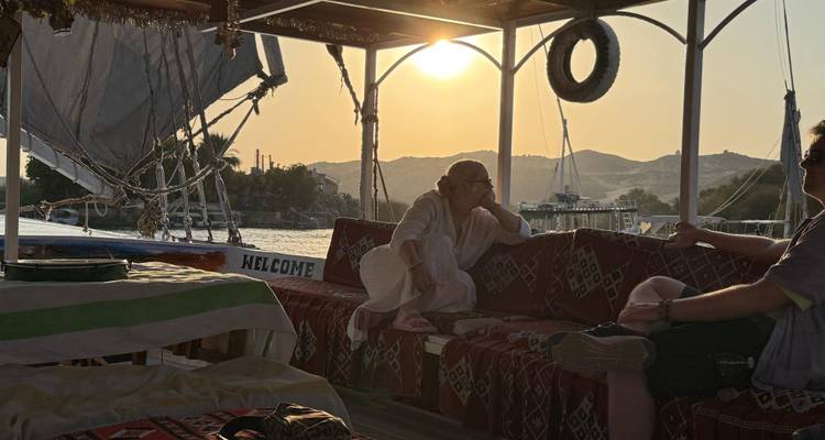 Deux voyageurs se détendent sur des bancs rembourrés à bord d'une felouque traditionnelle tandis que le soleil se couche sur le Nil, projetant une lumière chaleureuse sur les collines lointaines.
