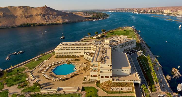 Vue aérienne du complexe hôtelier Mövenpick sur une île du Nil, avec une piscine turquoise, des terrasses bordées de palmiers et des collines désertiques environnantes.