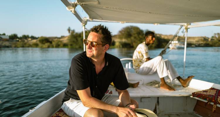 Traveller relaxes on the bow of a small white felucca while the captain navigates the calm Nile waters.