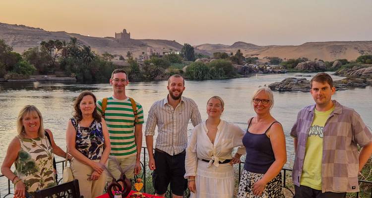 Small tour group smiles for a photo beside the Nile at dusk with desert hills in the distance.