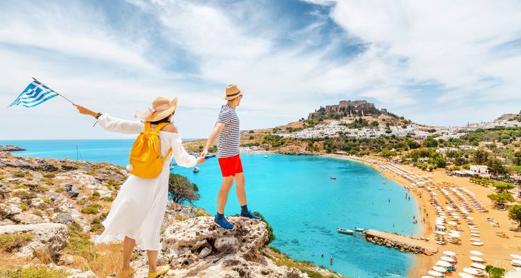 Couple with Greek flag overlooking a beach and ancient ruins.