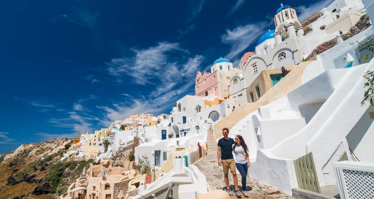 Two people walking on a narrow path in front of white and blue buildings on a cliff.