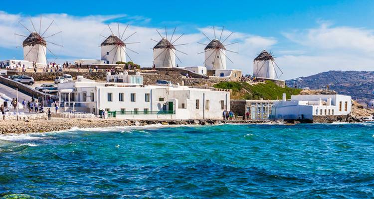 Row of iconic Mykonos windmills overlooking the Aegean Sea with white buildings along rocky shore.