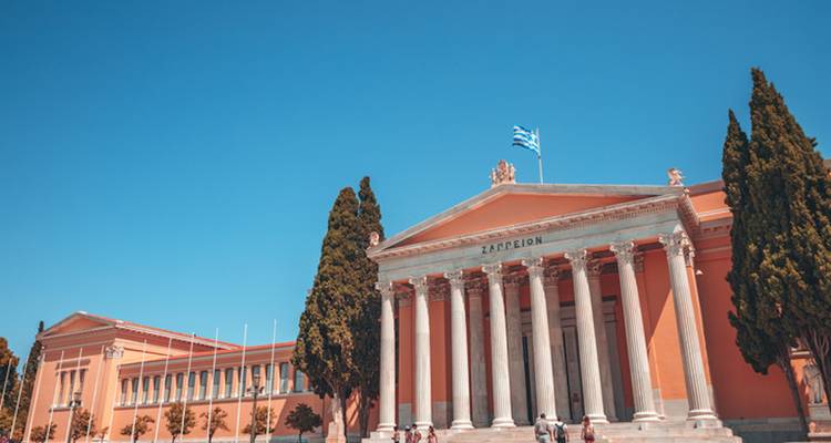 Classical Zappeion Hall with Greek flag flying above colonnade against clear blue sky.