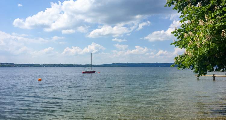 Un lago tranquilo salpicado con un velero solitario bajo un cielo parcialmente nublado, enmarcado por ramas frondosas.