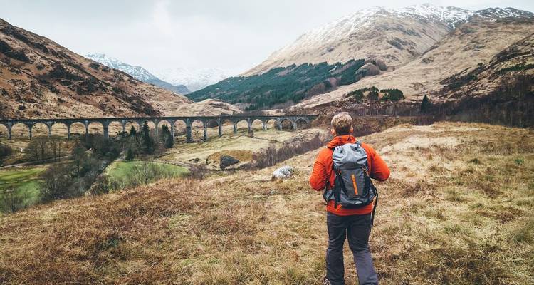 Man die een oranje jas draagt wandelt in een heuvelachtig landschap met een viaduct.