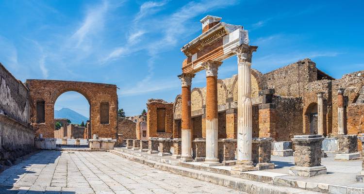 Des ruines ensoleillées et des colonnes dressées de l'ancienne Pompéi bordent un large forum pavé sous un ciel bleu vibrant.