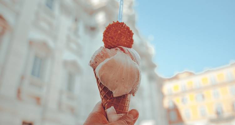 Gros plan d'une main tenant un cornet de gelato crémeux contre une place européenne ensoleillée floue.